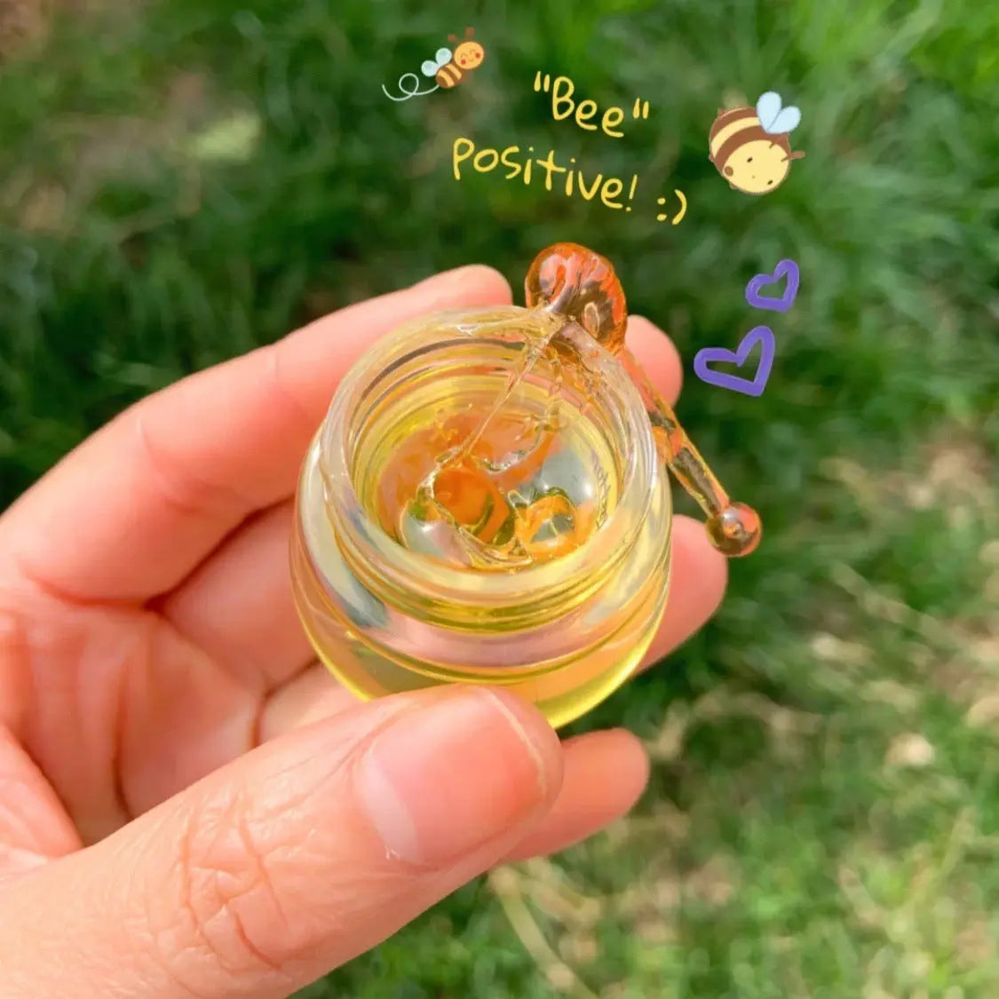 Hand holding a small honey jar with a spoon, surrounded by cheerful bee illustrations, outdoors.